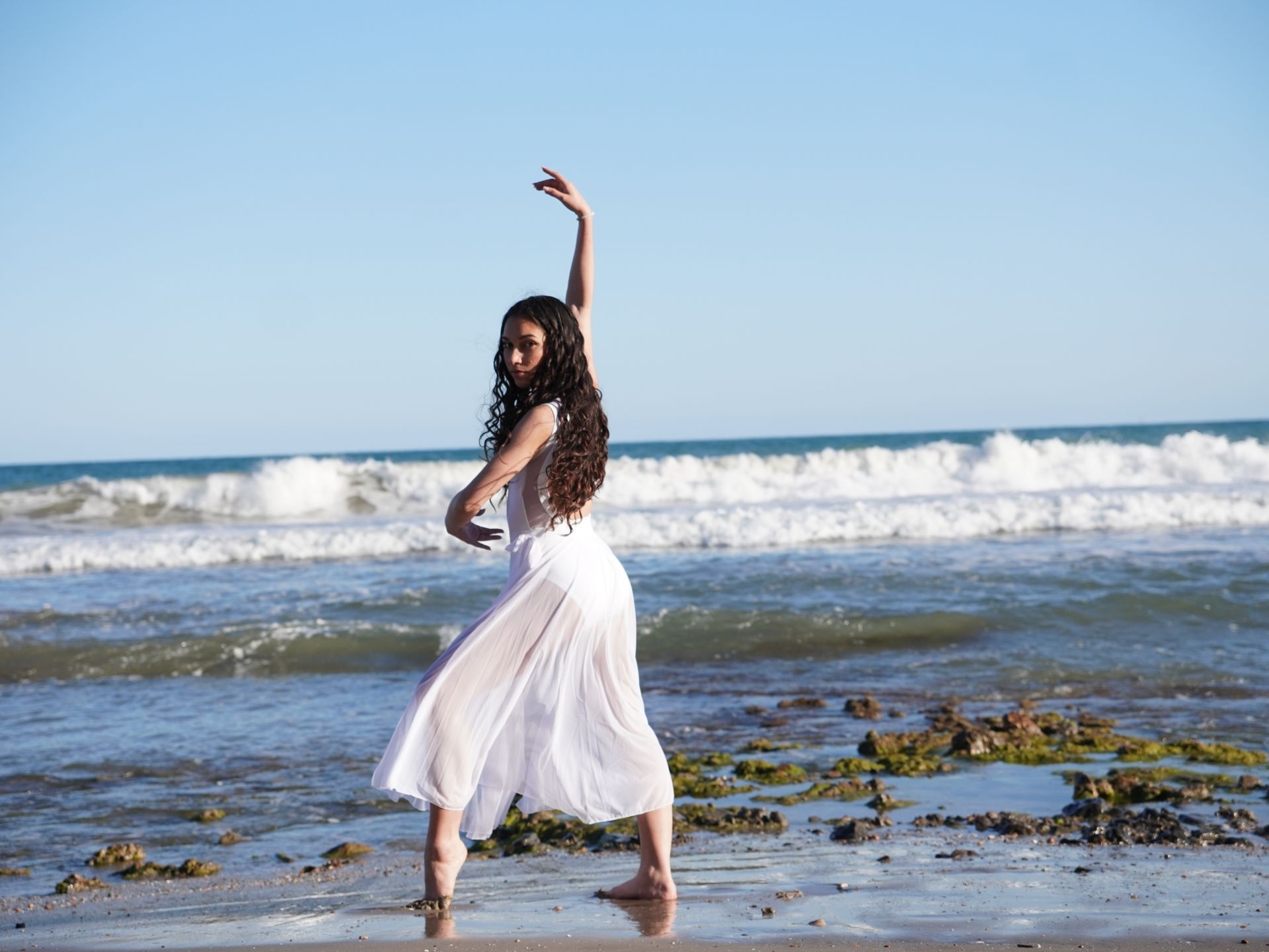 Mujer con vestido blanco bailando en una playa con olas y cielo azul de fondo.