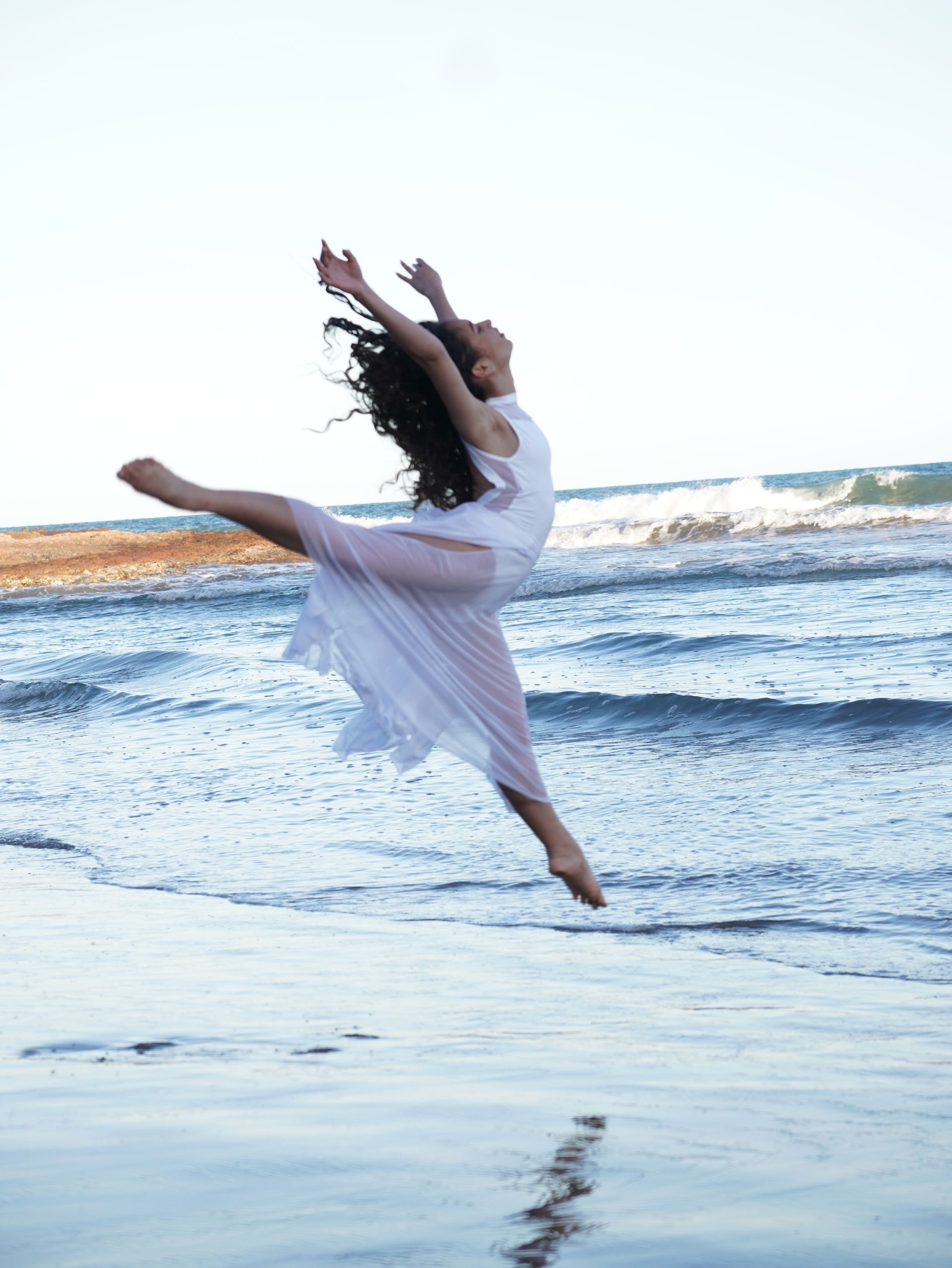 Bailarina con vestido blanco saltando sobre las olas del océano en la playa.