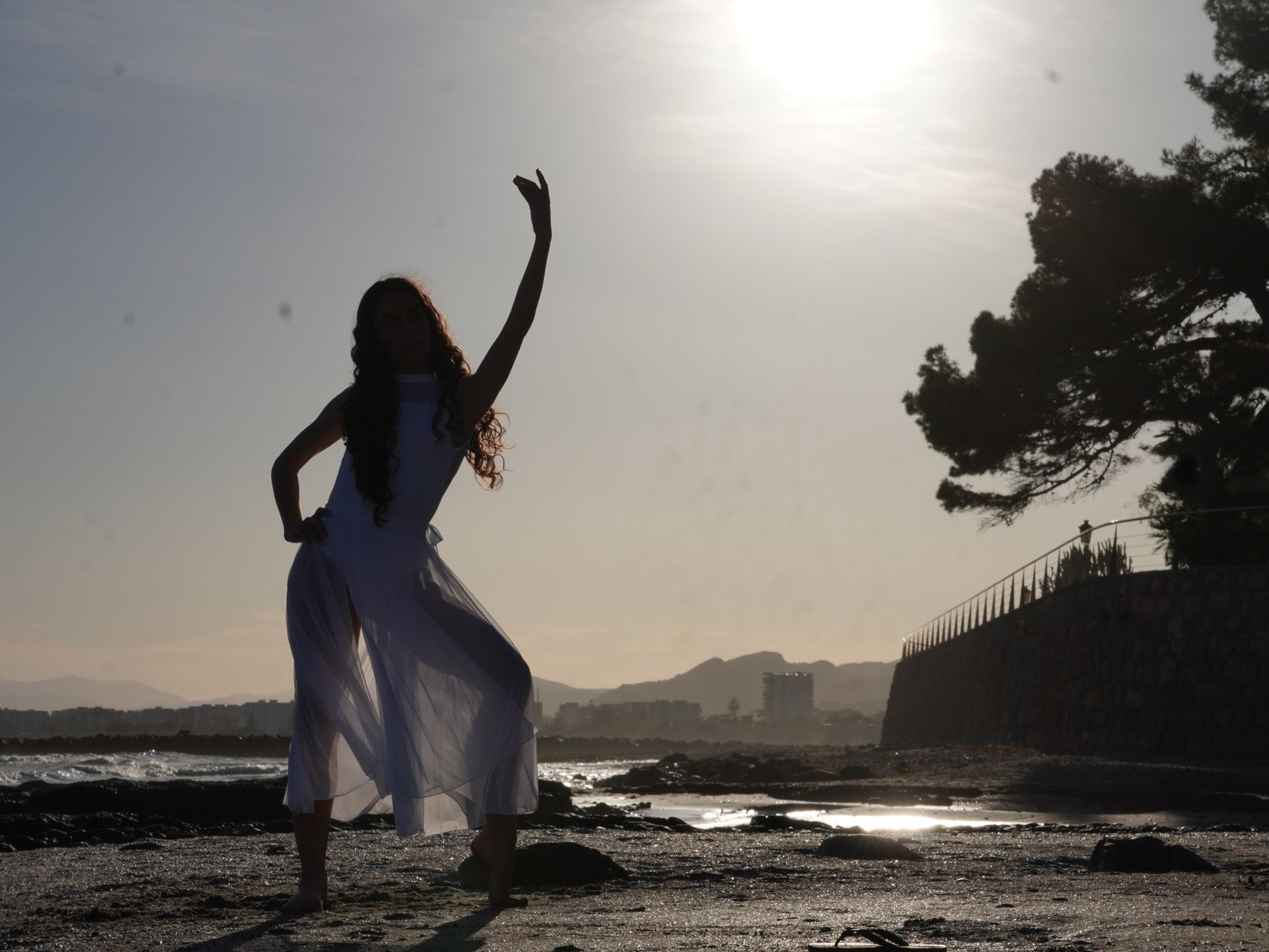 Silueta de una persona bailando en una playa al atardecer, con un cielo brillante y una costa bordeada de árboles.