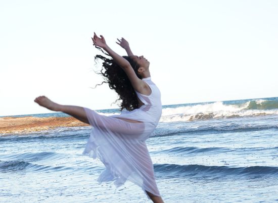 Bailarina con vestido blanco saltando en la playa con los brazos en alto, con las olas del océano de fondo.