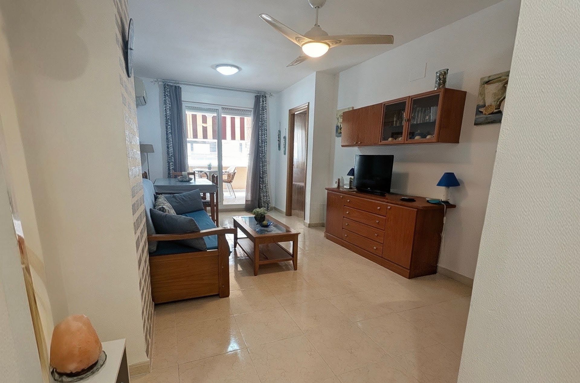 Sala de estar luminosa con sofá, mueble para TV, ventilador de techo y luz de balcón al fondo.