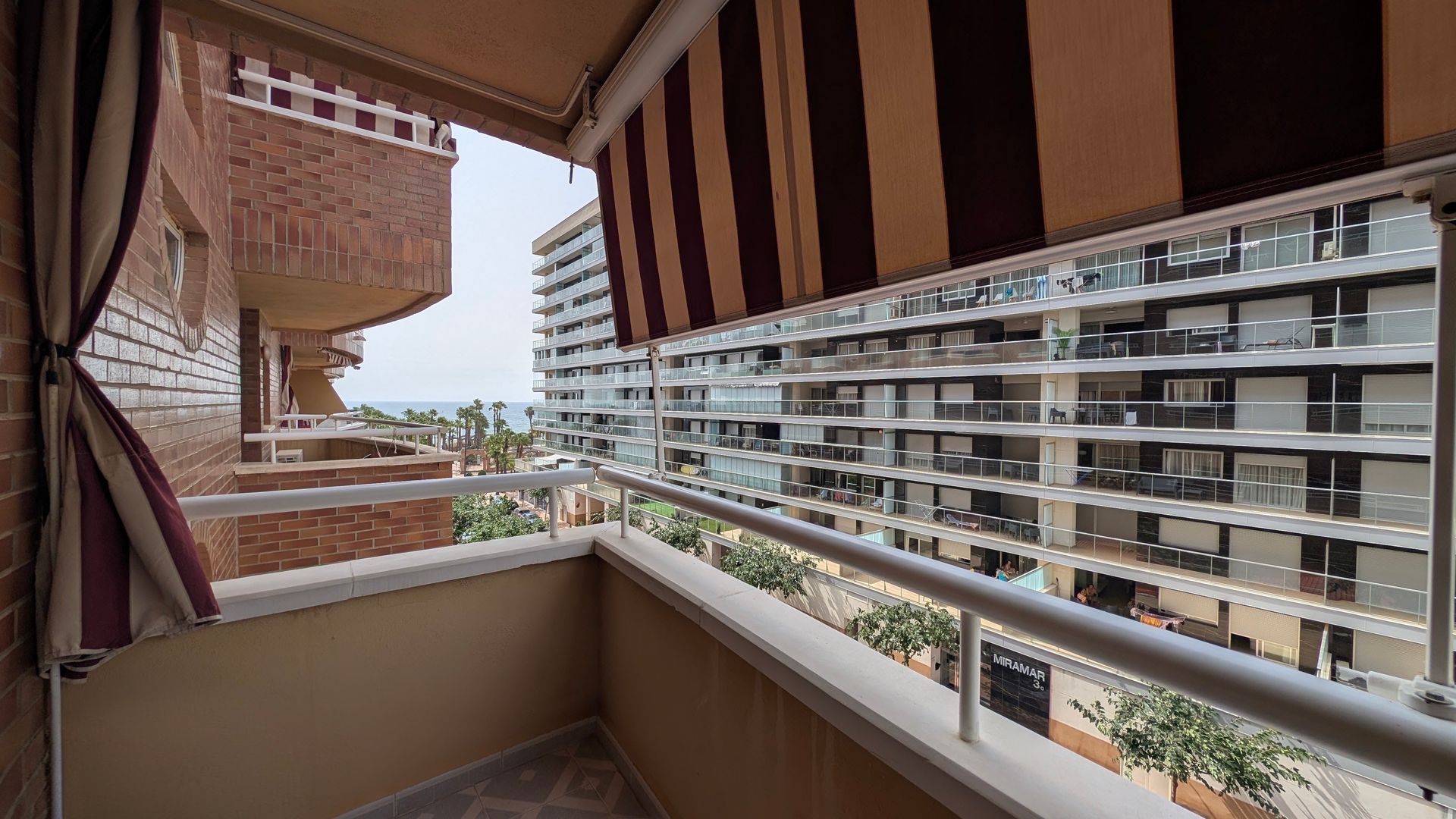 Vista desde el balcón con toldo a rayas, barandilla blanca y edificios de apartamentos al otro lado de la calle.