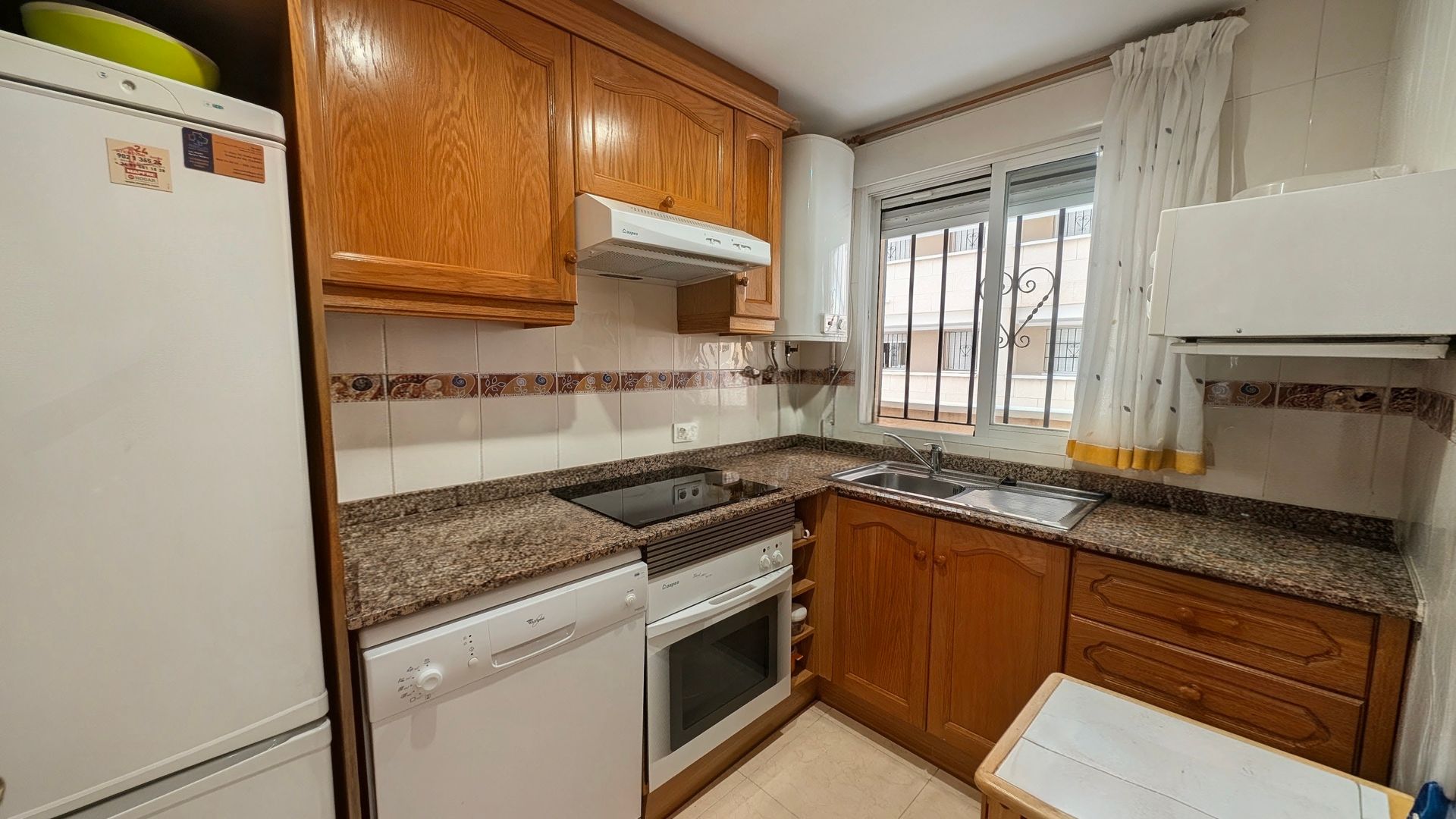 Cocina compacta con armarios de madera, encimeras de granito, electrodomésticos blancos y una ventana sobre el fregadero.