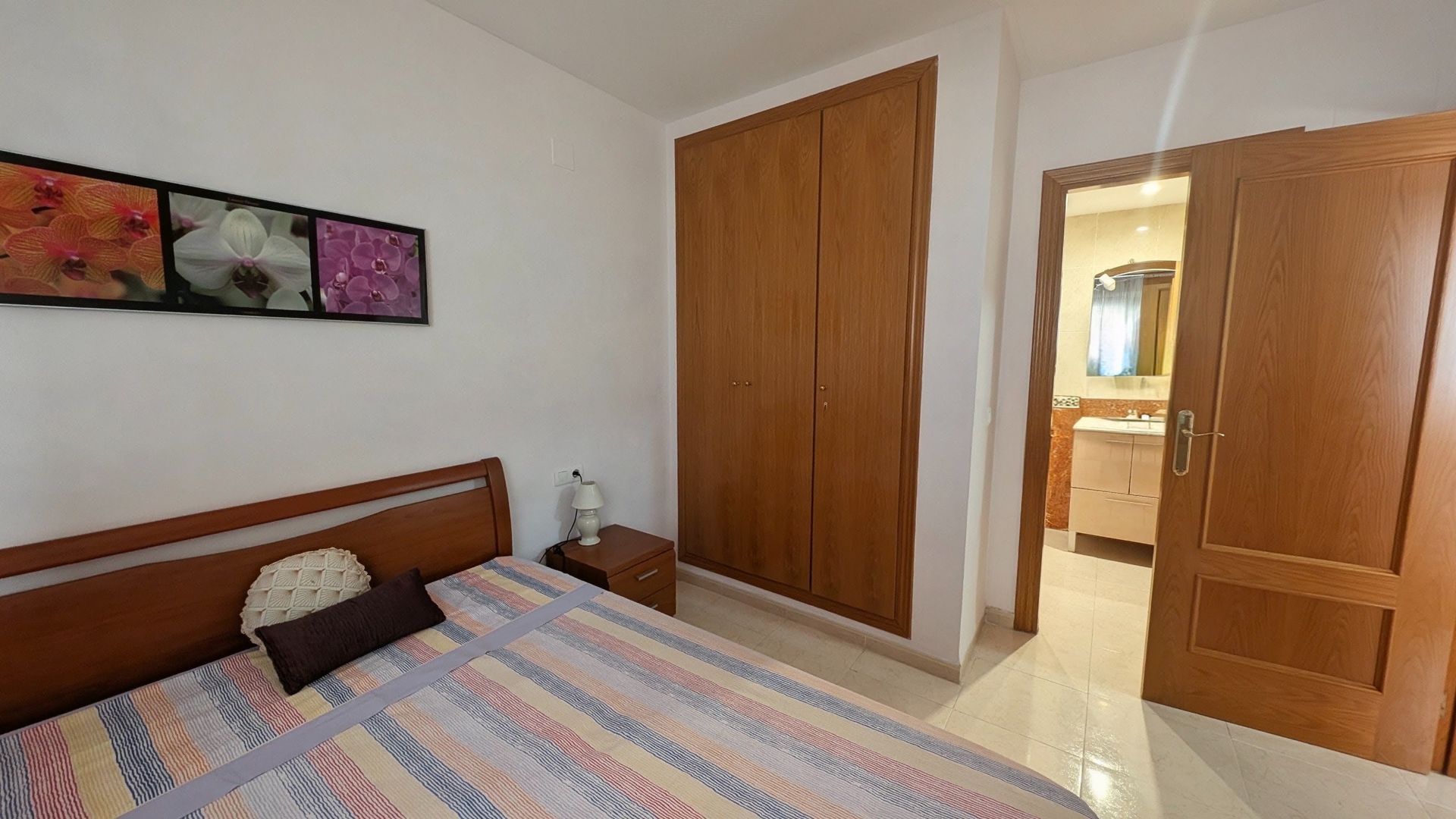 Dormitorio con cama de madera, colcha estampada, cuadros en la pared, armario y puerta abierta al baño.