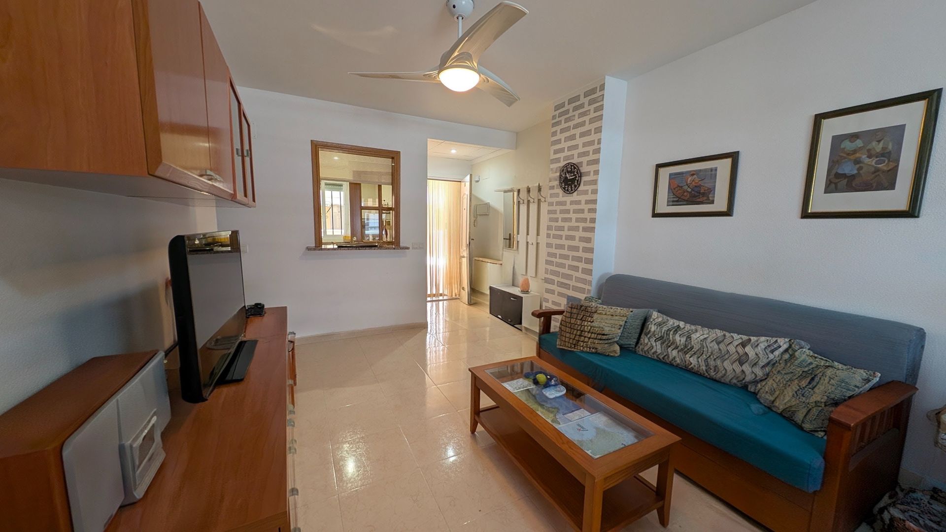 Sala de estar luminosa con sofá azul, mesa de centro de madera, cuadros en la pared, televisor y zona de cocina abierta.