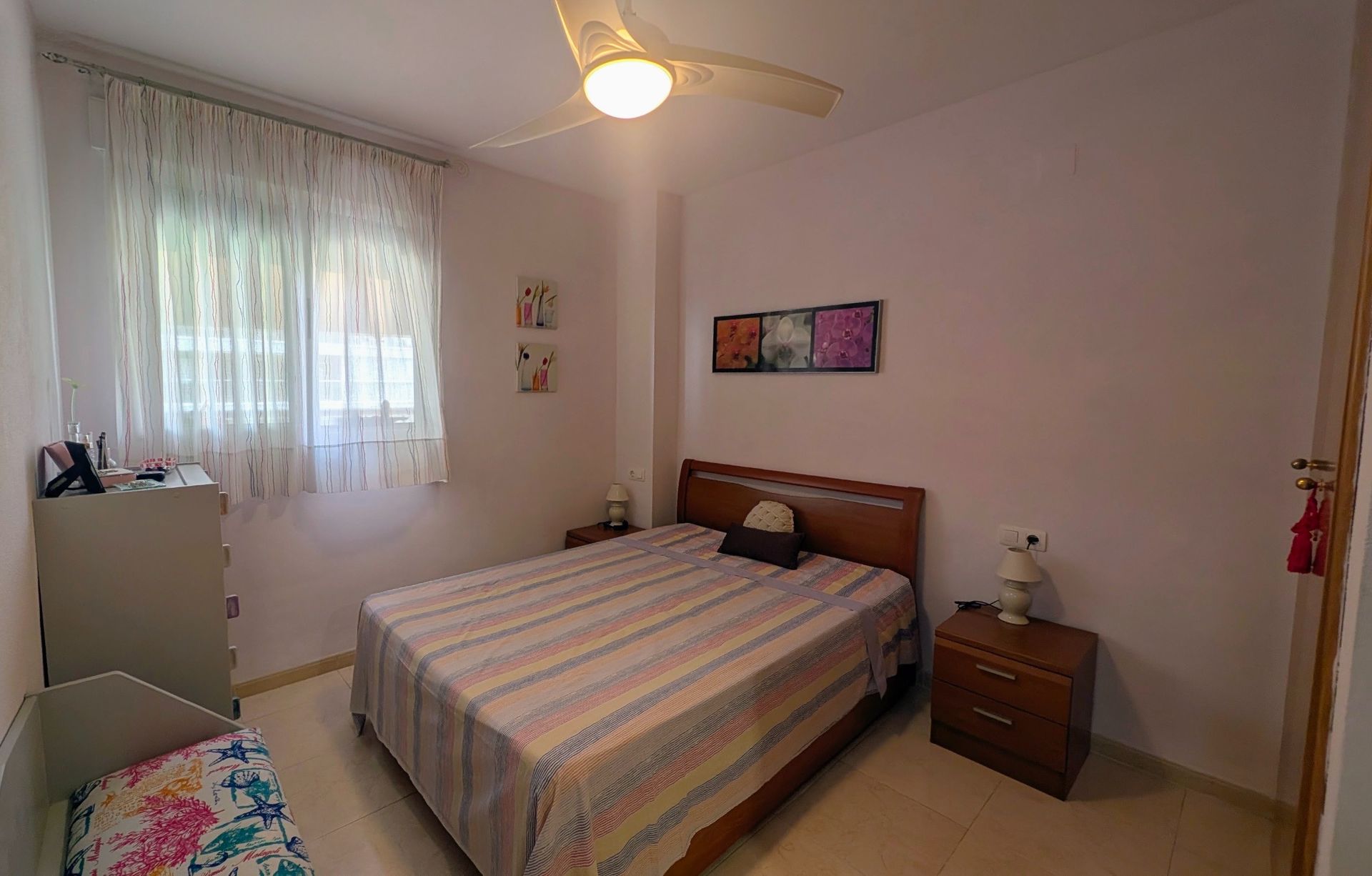 Habitación luminosa con cama, mesitas de noche, ventana, ventilador de techo y cómoda junto a una pared blanca.