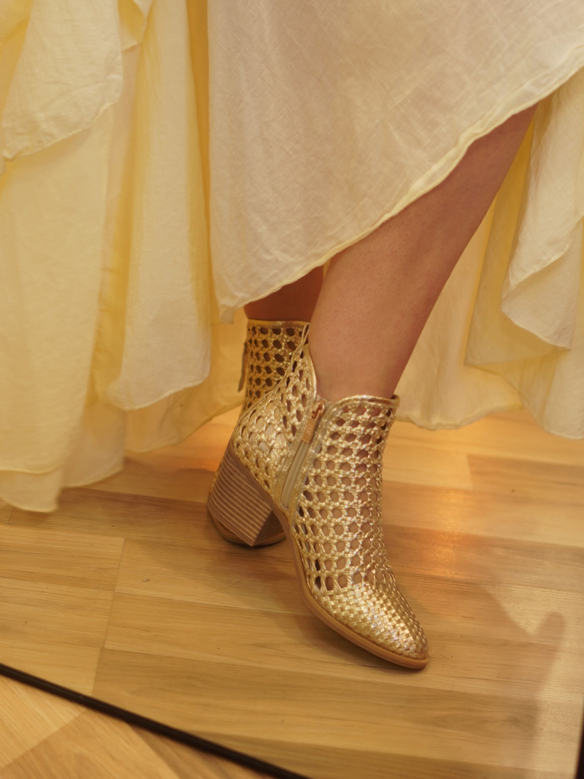Botines dorados tejidos bajo un vestido vaporoso color crema sobre un suelo de madera.