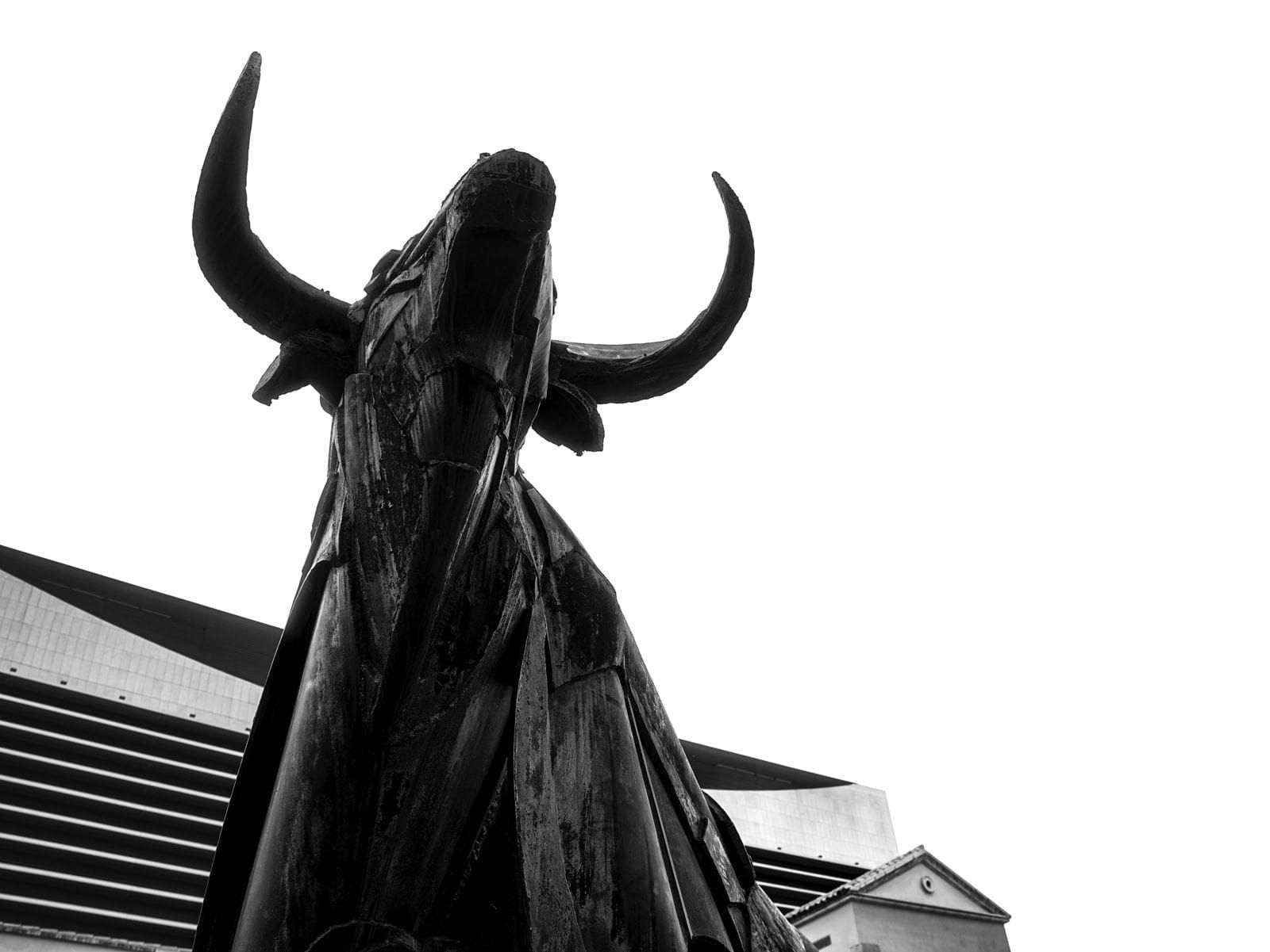 Estatua oscura con cuernos recortada contra un edificio moderno y un cielo nublado.