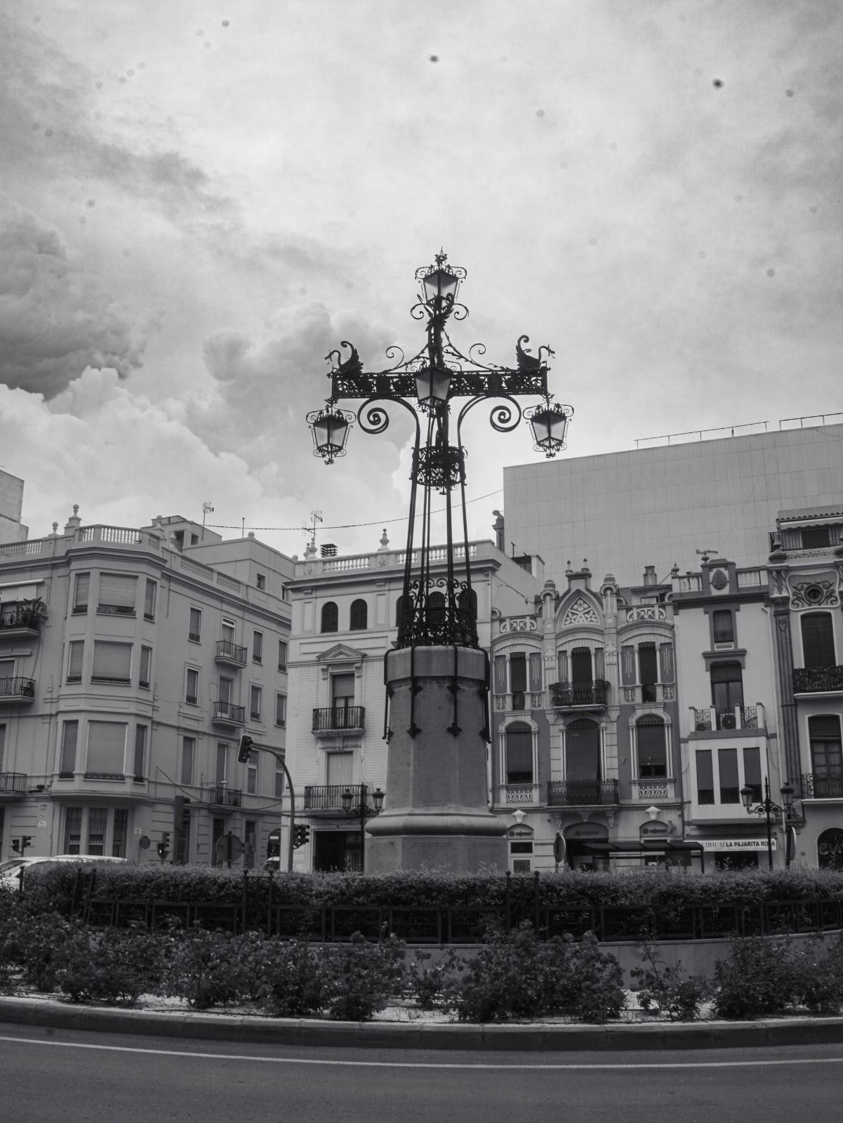Plaza de la ciudad en blanco y negro con una farola ornamentada y edificios históricos bajo un cielo nublado.