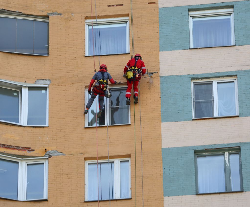 Dos rescatistas vestidos con trajes rojos permanecen de pie junto a la ventana de un edificio de apartamentos de ladrillo.