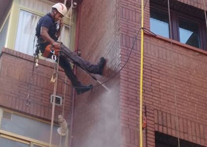 Un trabajador, suspendido por cuerdas, limpia un edificio de ladrillos con una hidrolavadora.
