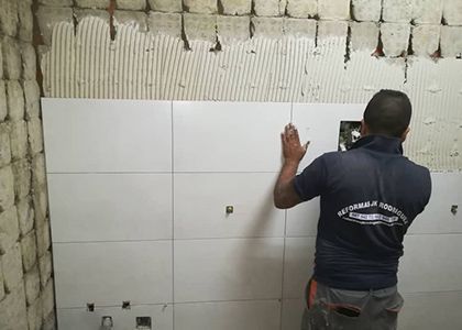 Obrero instalando azulejos blancos en una pared interior, con yeso sin terminar y cableado expuesto en la parte superior.