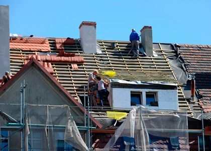 Obreros reparando el techo de una casa bajo un cielo azul, con andamios y tejas rojas cerca.