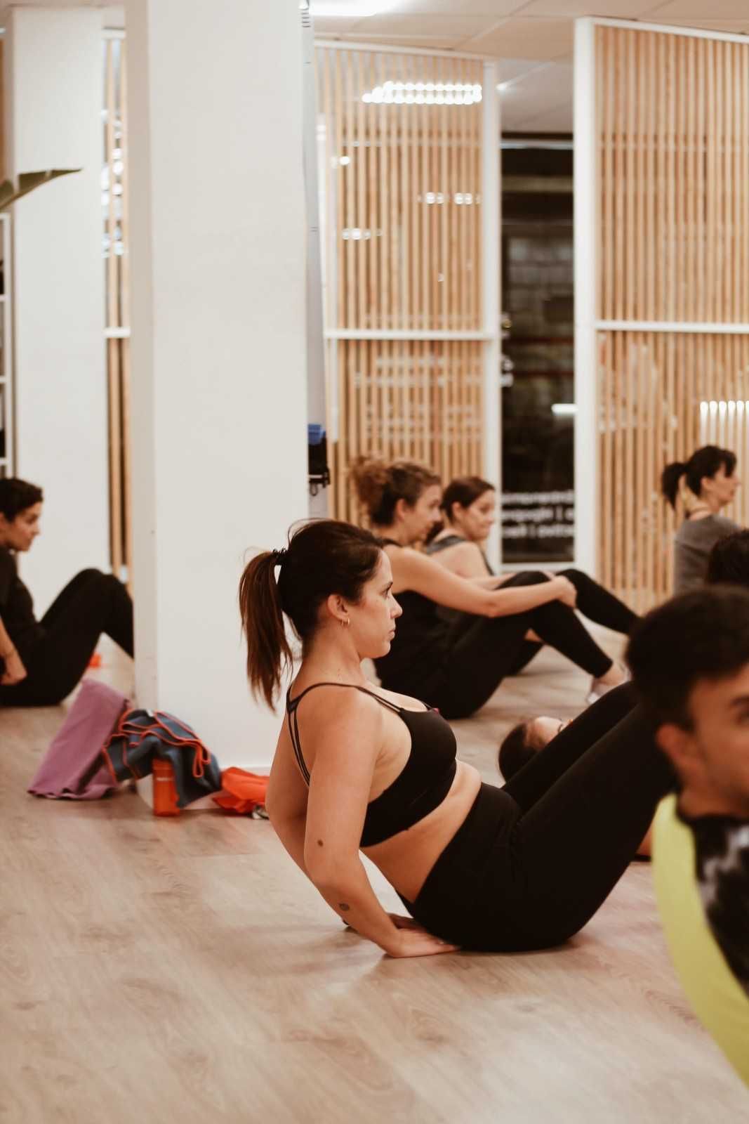 Personas haciendo ejercicio sobre colchonetas en un estudio luminoso, realizando ejercicios abdominales sentados.