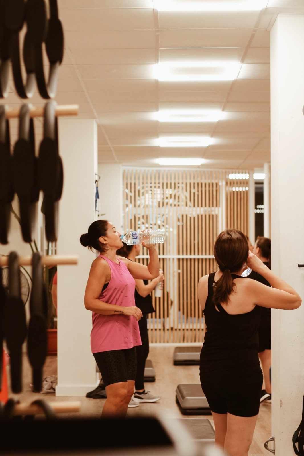 Dos mujeres con ropa deportiva bebiendo agua en el pasillo de un gimnasio junto a una pared llena de pesas.