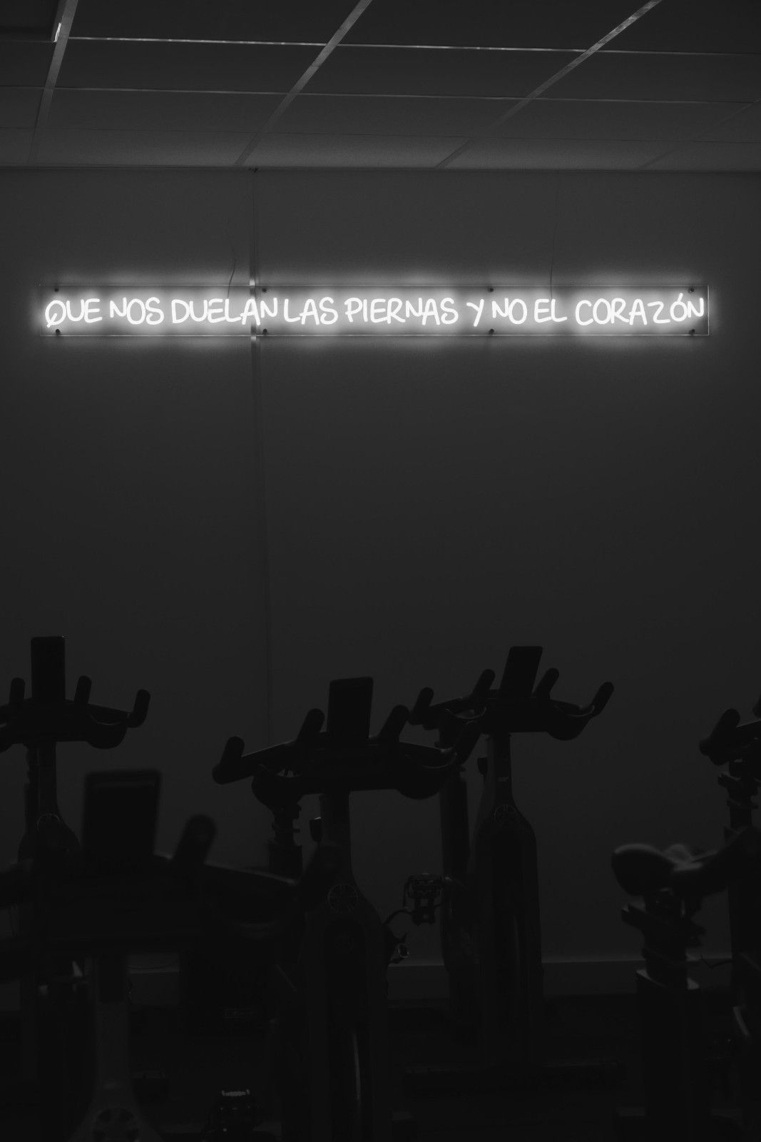 Gimnasio con poca luz, con siluetas de máquinas de ejercicio y un letrero de neón brillante en la pared.