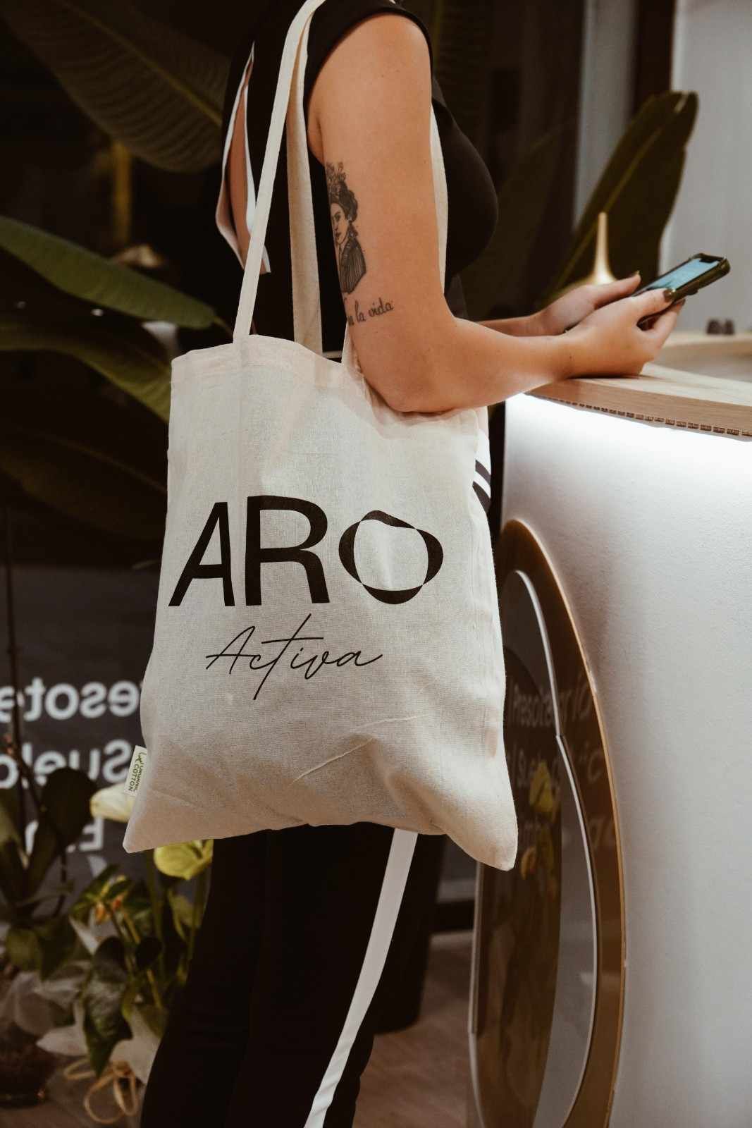 Una persona sostiene un teléfono junto a una ventana y lleva una bolsa de tela blanca de la marca ARO sobre ropa negra.