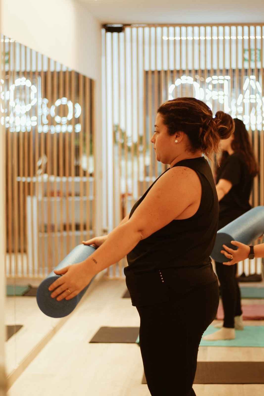 Mujer sosteniendo una pelota de ejercicio azul en un gimnasio luminoso, con espejos y otra persona al fondo.