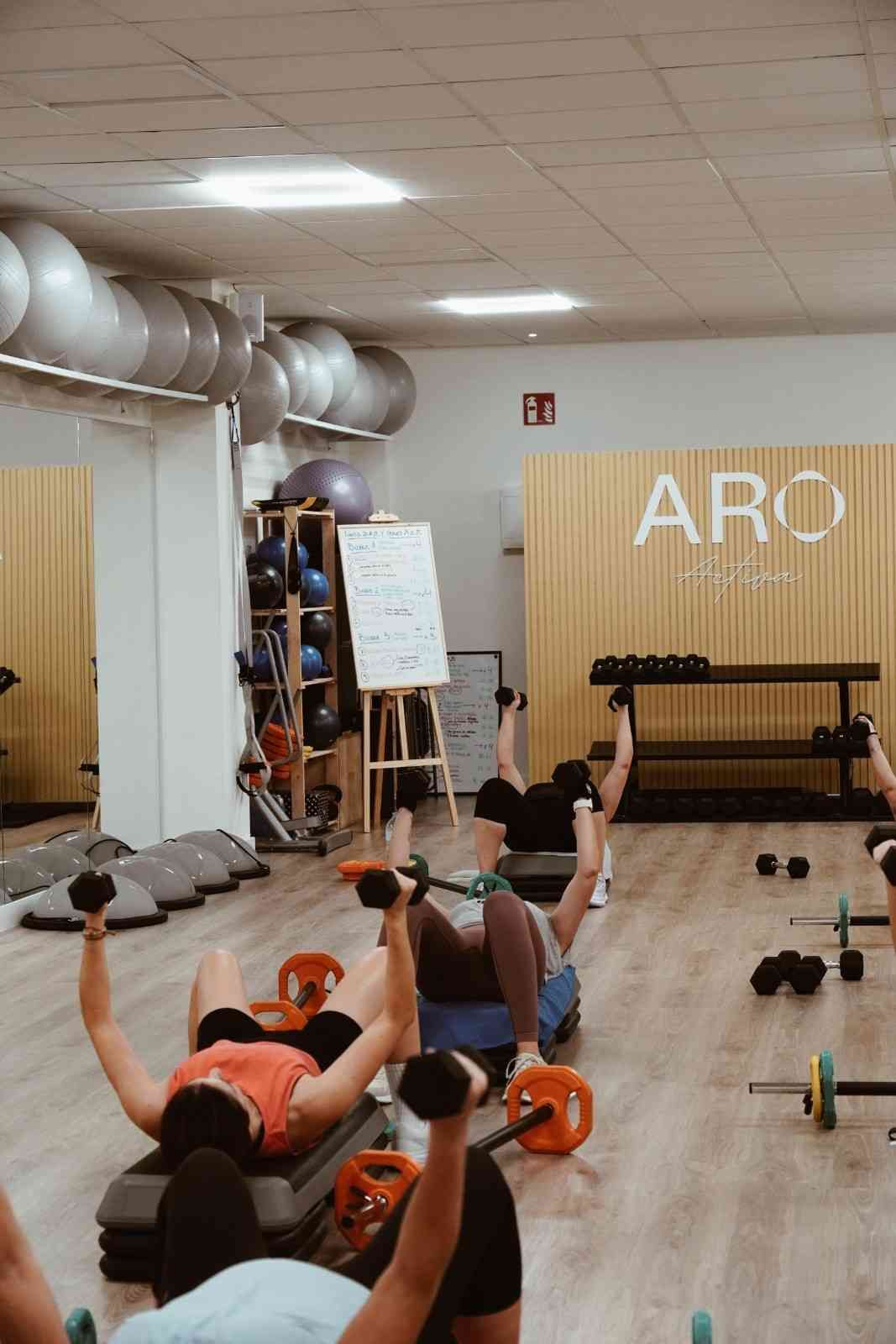 Personas realizando ejercicios de suelo con mancuernas en un gimnasio con paredes de madera y espejos.