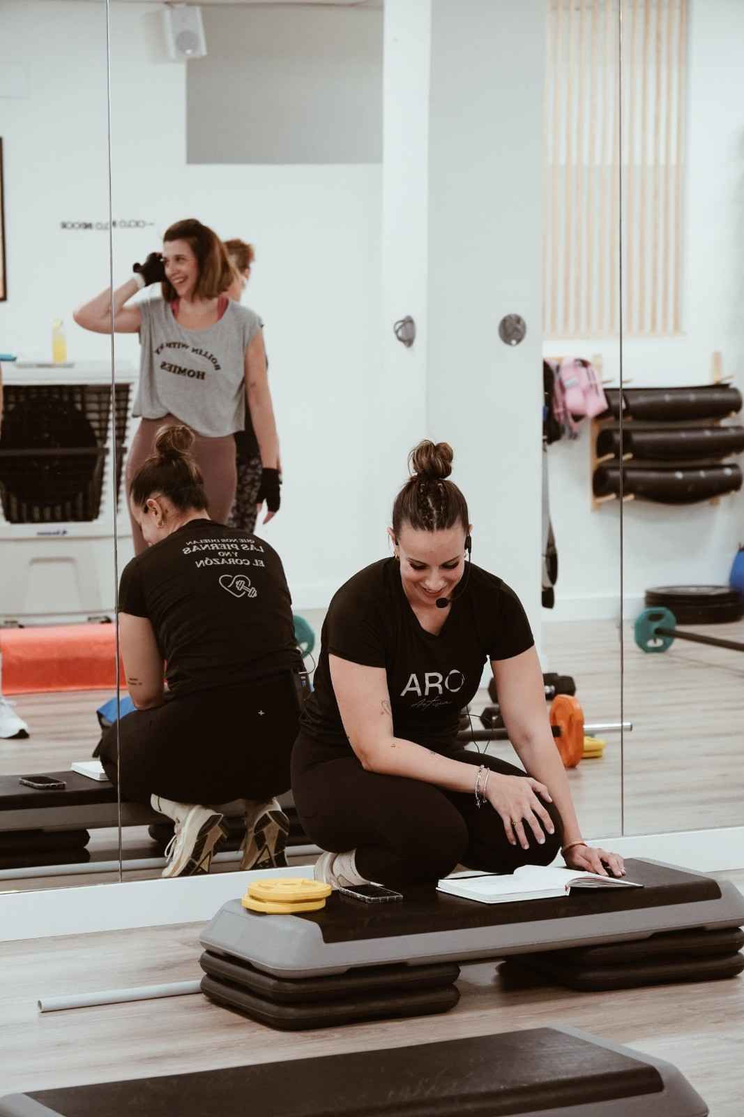 Dos mujeres haciendo ejercicio en un estudio con espejos; una está arrodillada sobre colchonetas.