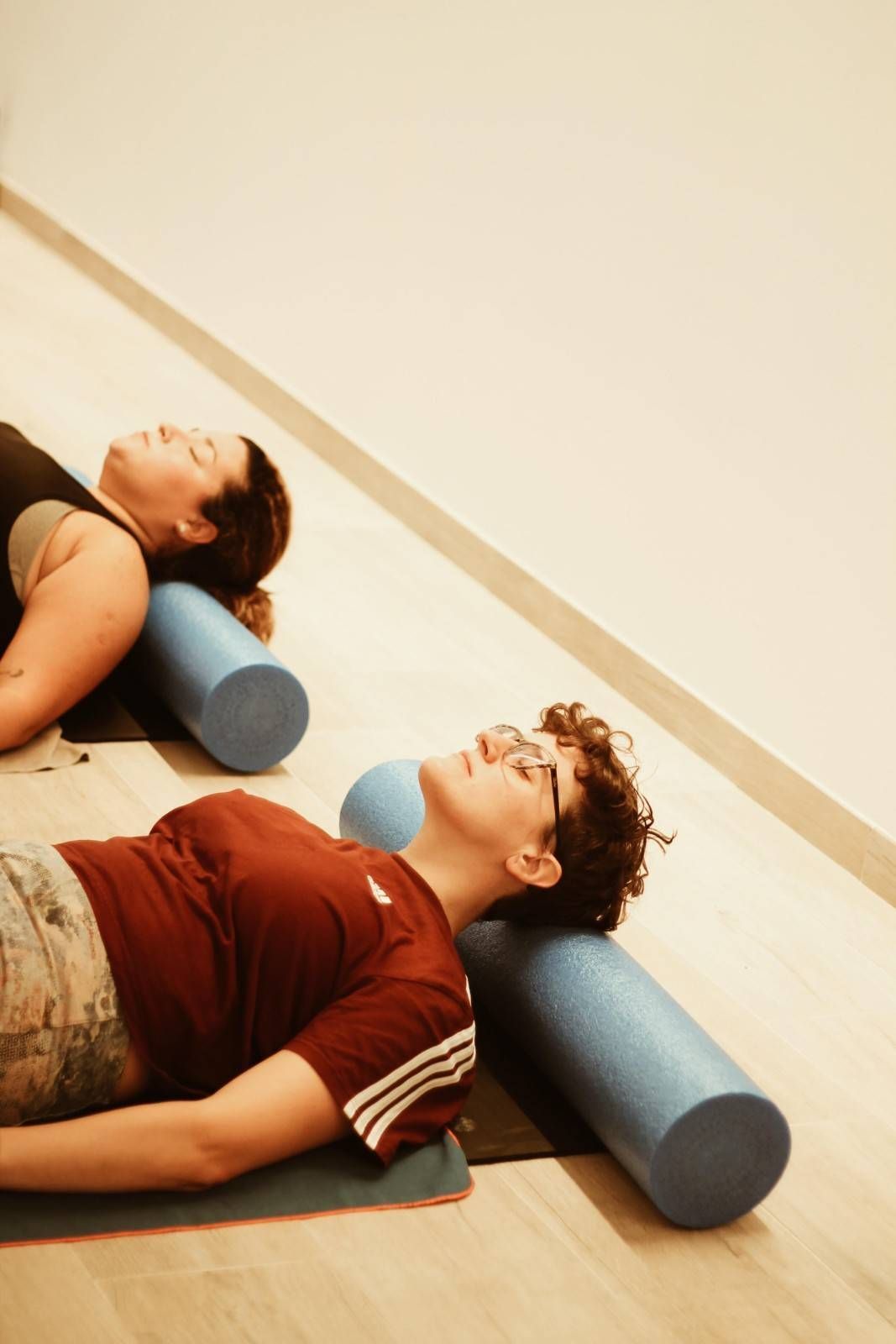 Dos personas tumbadas sobre esterillas de yoga usando rodillos de espuma en un estudio luminoso.