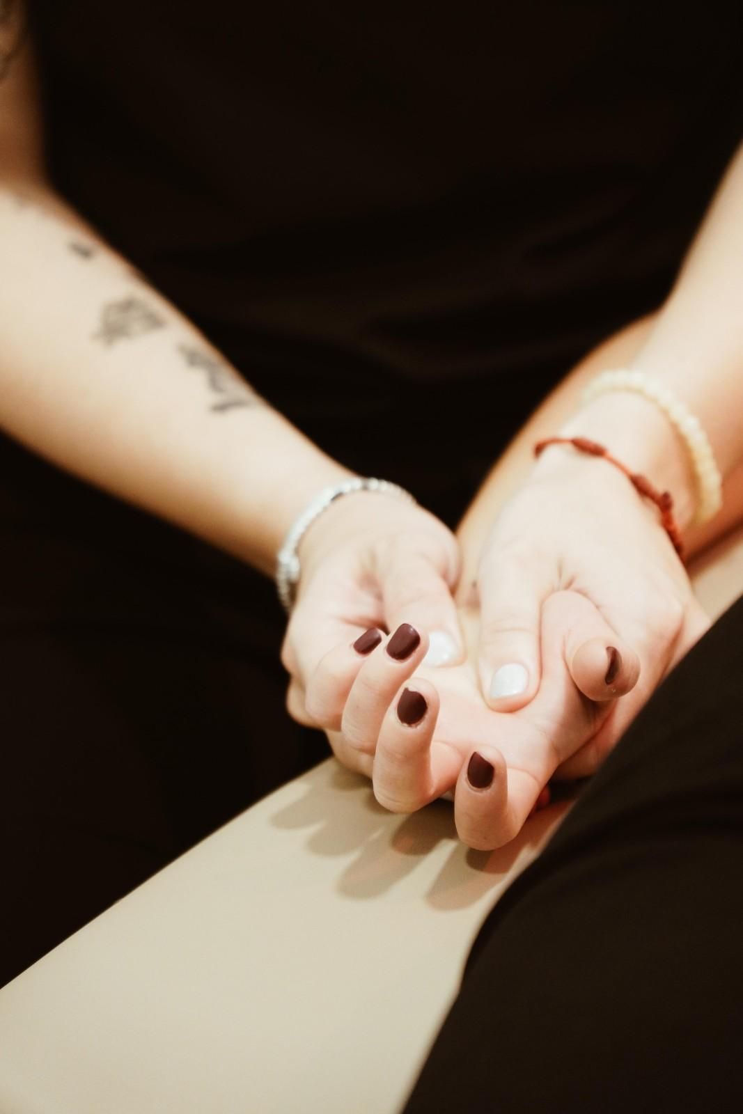 Dos personas tomadas de la mano, una con tatuajes y pulseras, sentadas muy cerca una de la otra en un ambiente con poca luz.