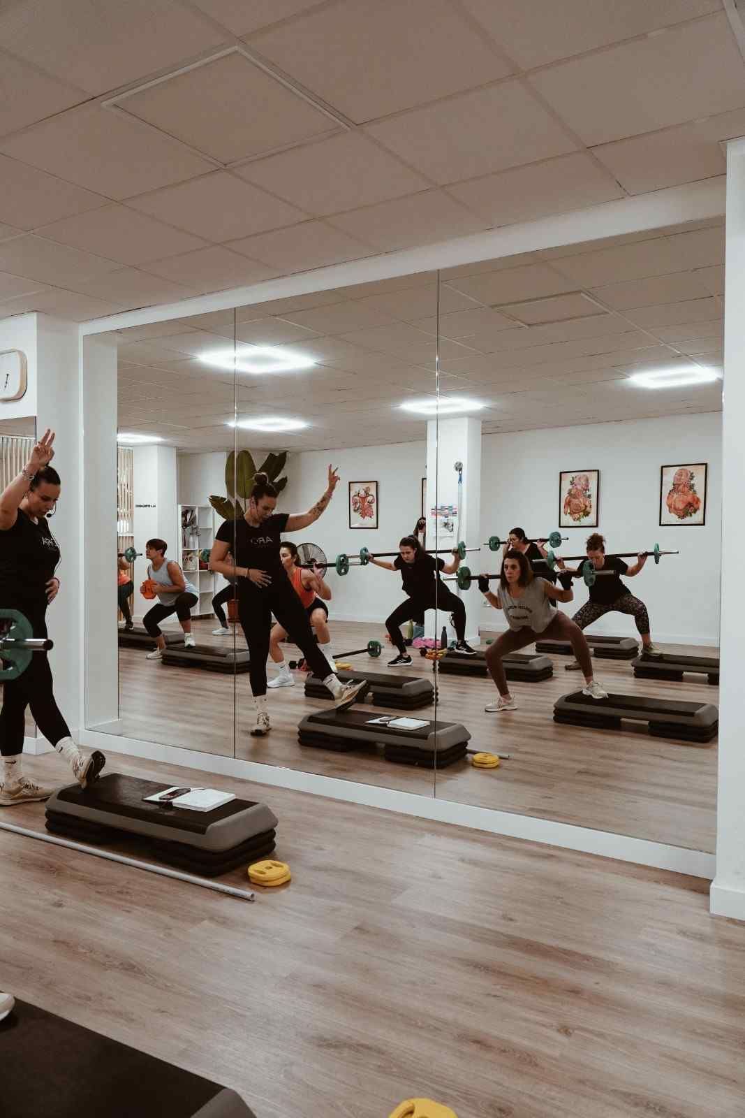 Personas haciendo ejercicio en un gimnasio con espejos, plataformas escalonadas y suelo de madera.
