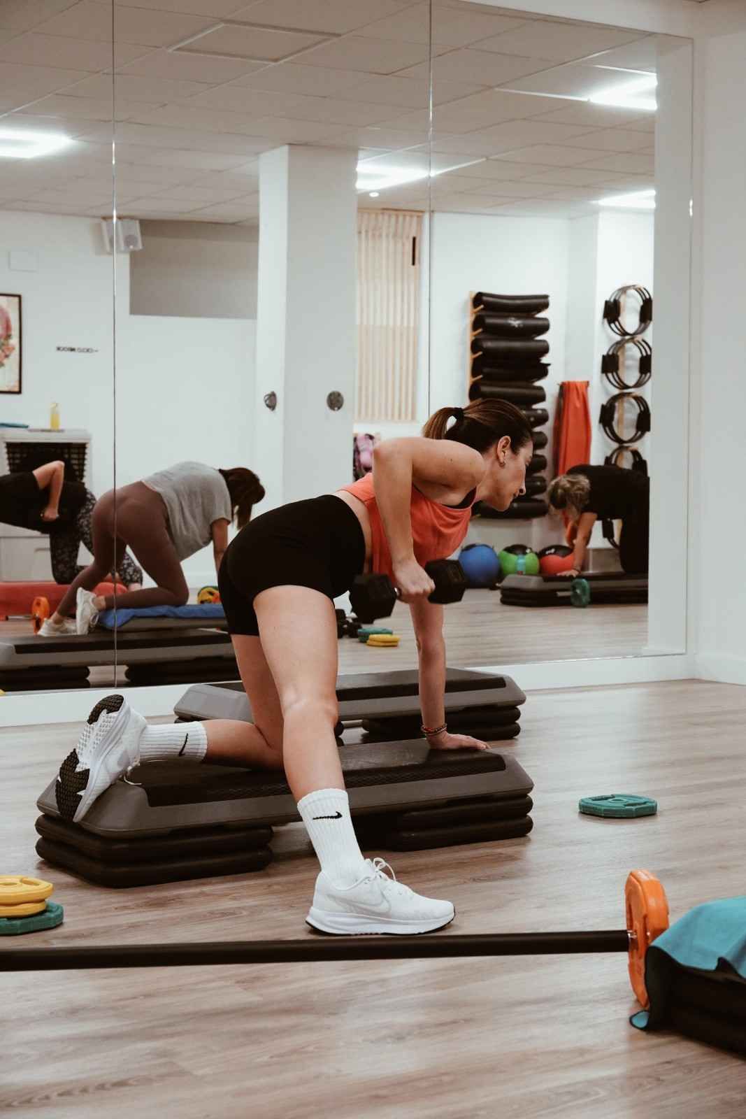 Mujer estirándose sobre una plataforma con escalones en un gimnasio, reflejada en un espejo.