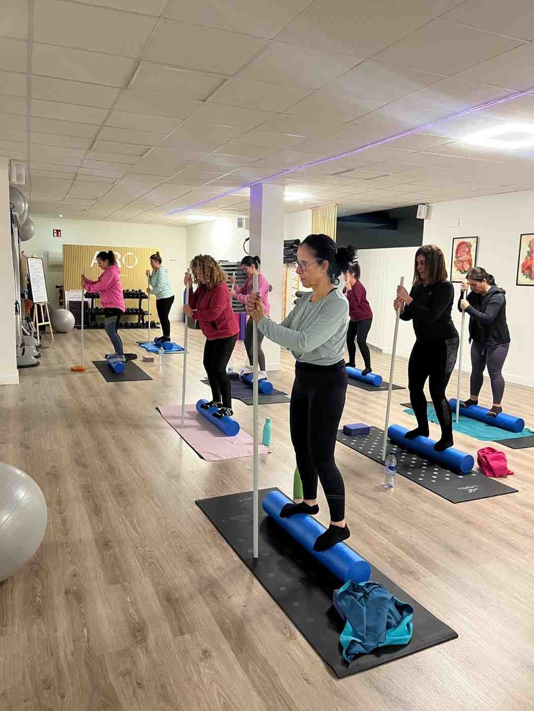 Clase de fitness en grupo en un estudio con espejos, donde la gente mantiene el equilibrio sobre escalones azules.
