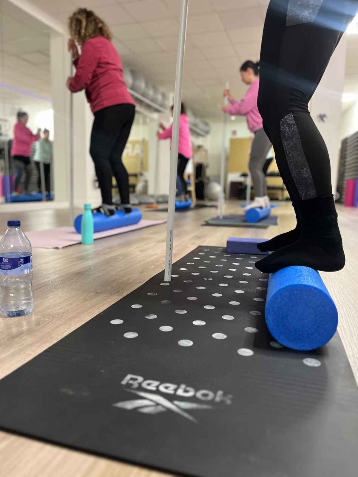 Personas manteniendo el equilibrio sobre rodillos de espuma en un estudio con espejos durante una clase de fitness.