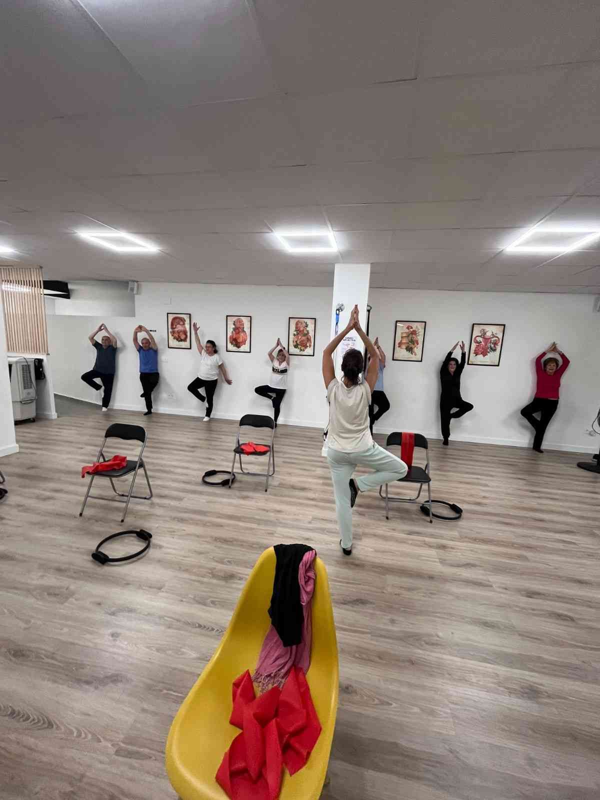 Personas realizando una postura de equilibrio de yoga en un estudio con sillas y esterillas en el suelo.