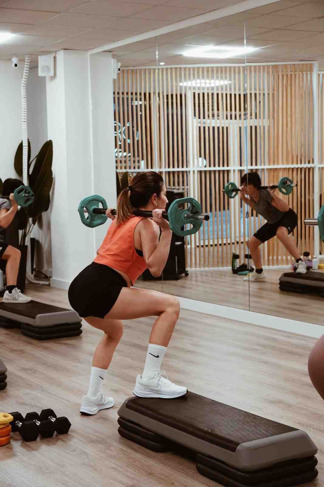 Mujer realizando ejercicios de subida de escalón con mancuernas en un gimnasio, vista a través de un espejo en la pared.