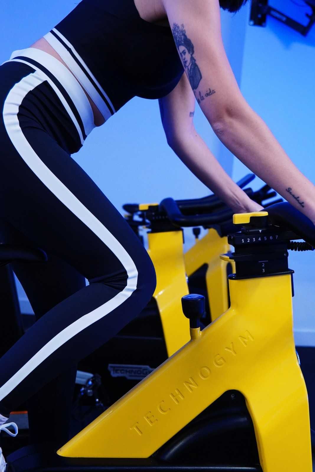 Persona montando una bicicleta estática amarilla en un gimnasio interior iluminado con luz azul.