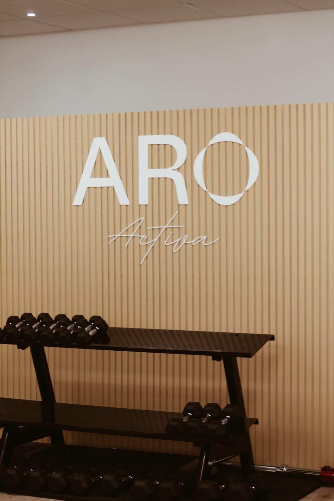 Letrero ARO en una pared de listones de madera clara sobre un estante para mancuernas en un gimnasio o estudio.