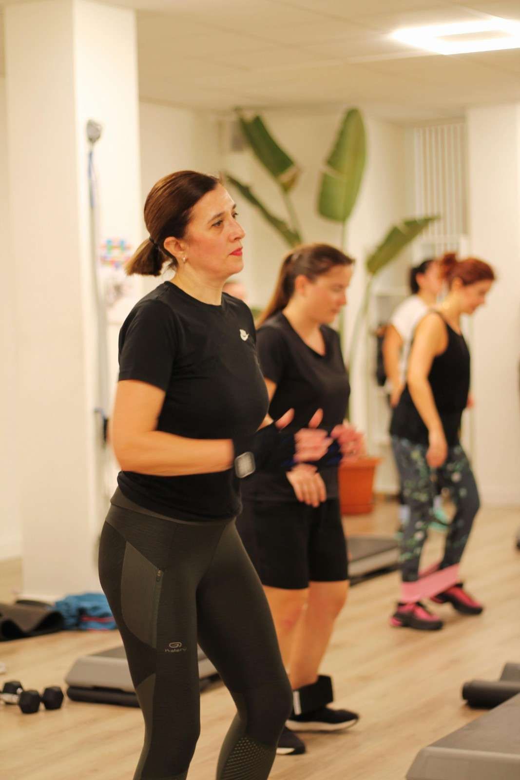Varias personas haciendo ejercicio en un estudio de danza con espejos; una mujer vestida de negro aparece en primer plano.