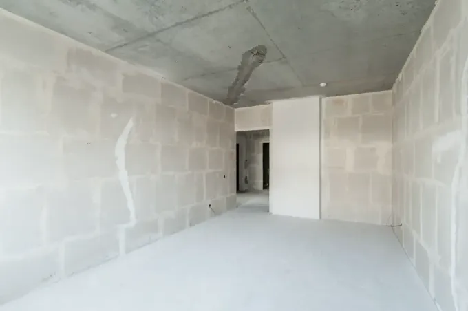 Habitación vacía en construcción con techo y paredes de hormigón, paneles de yeso blancos y una puerta.