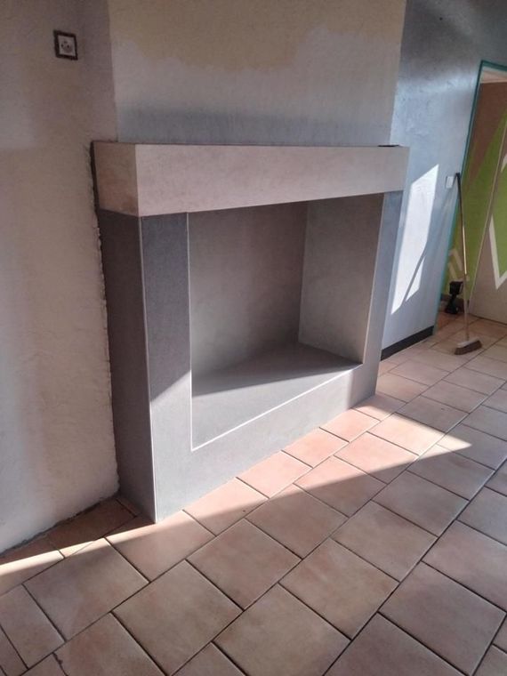 Alcôve de cheminée en béton avec un manteau beige clair dans une pièce intérieure carrelée, près d'une porte.