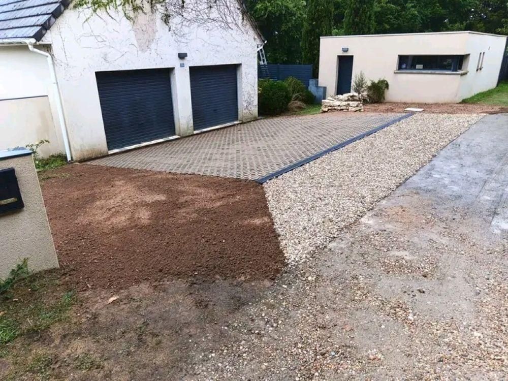 Allée menant à un garage avec deux portes sombres, chemin de gravier et maison aux murs blancs et petite terrasse.