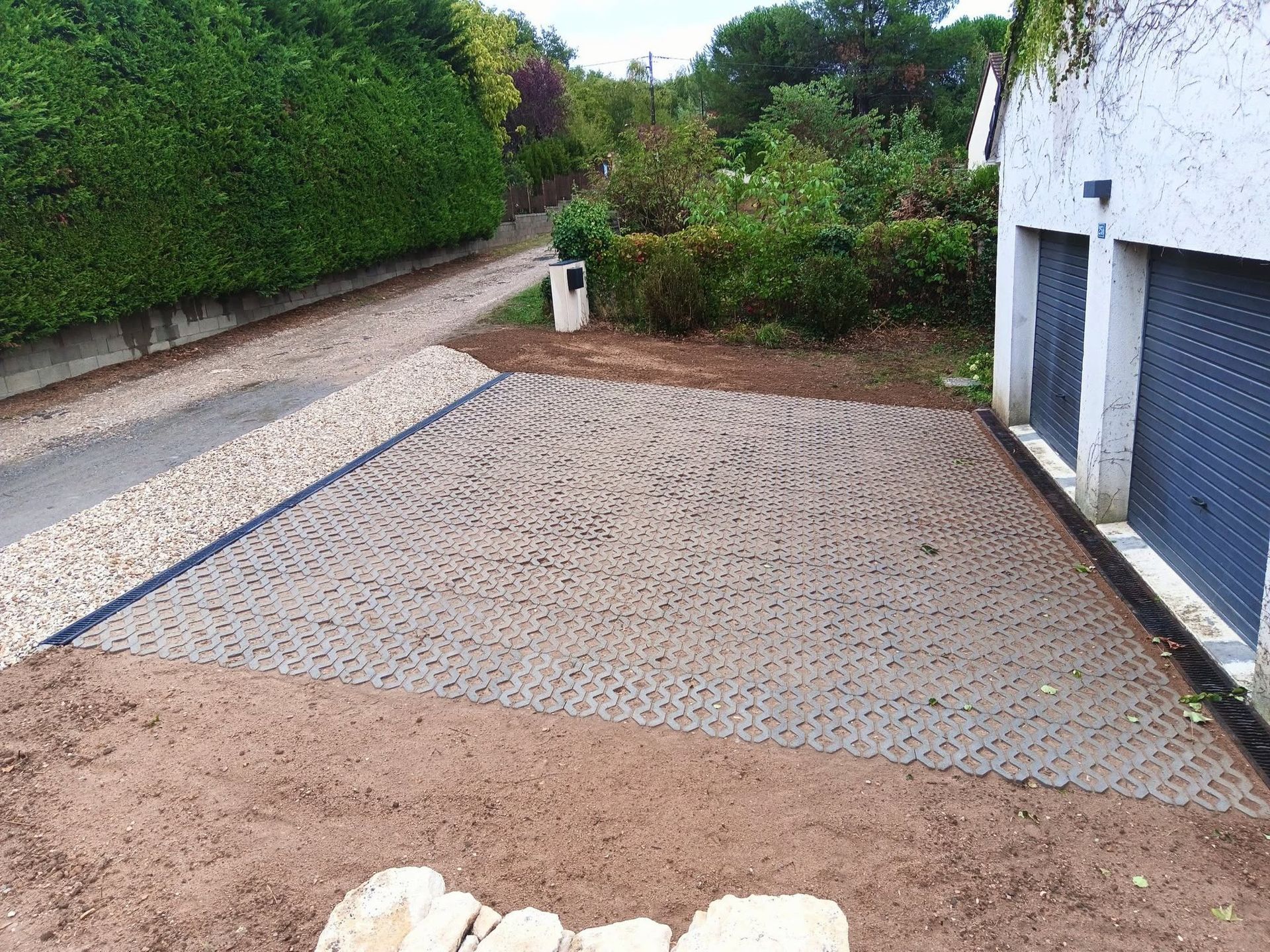Allée pavée longeant un garage blanc et des haies, avec un chemin de gravier et de la verdure du jardin au-delà.