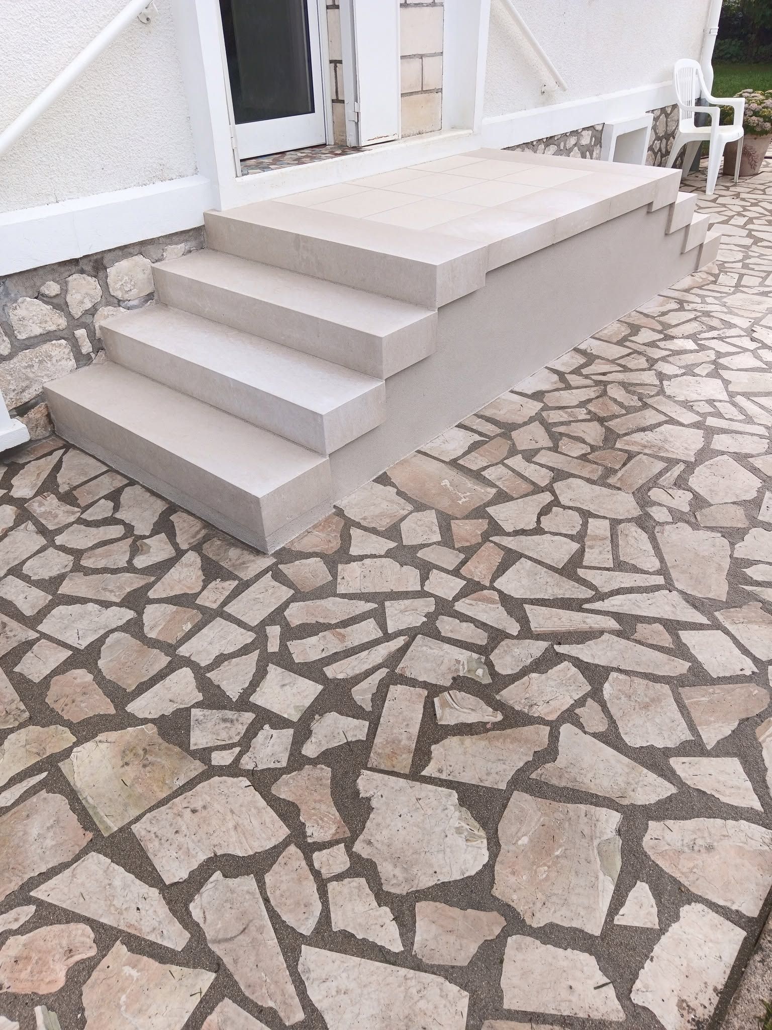 Des marches en pierre mènent à l'entrée d'un bâtiment blanc, donnant sur une terrasse en pierre à motifs.