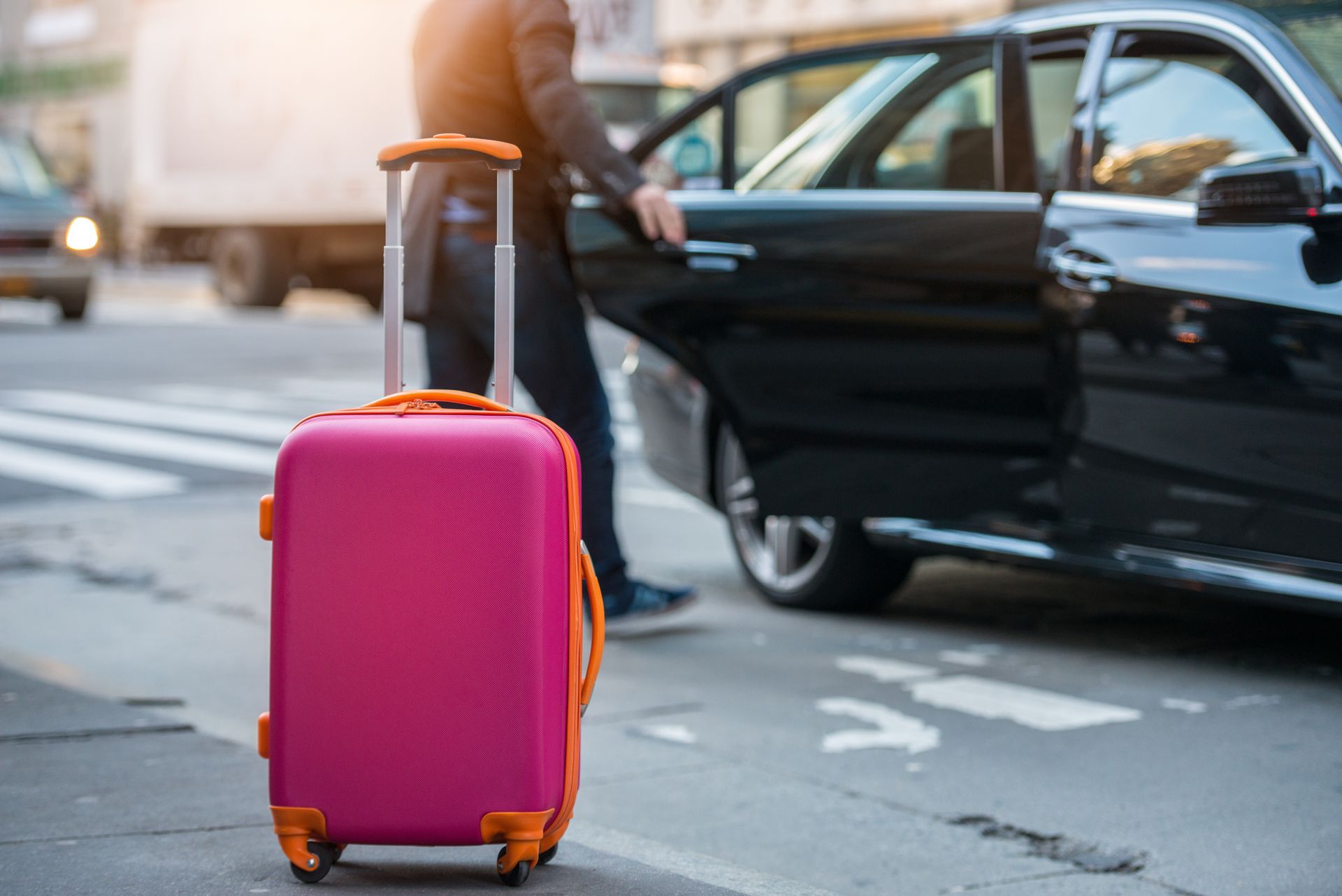Ein rosa Koffer neben einer Person, die auf einer Stadtstrasse die Tür eines schwarzen Autos öffnet.