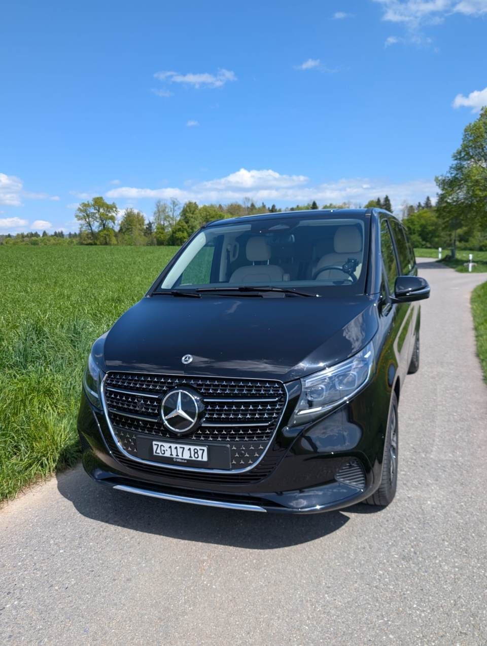 Ein schwarzer Mercedes-Transporter parkt auf einer Landstrasse neben grünen Feldern unter blauem Himmel.