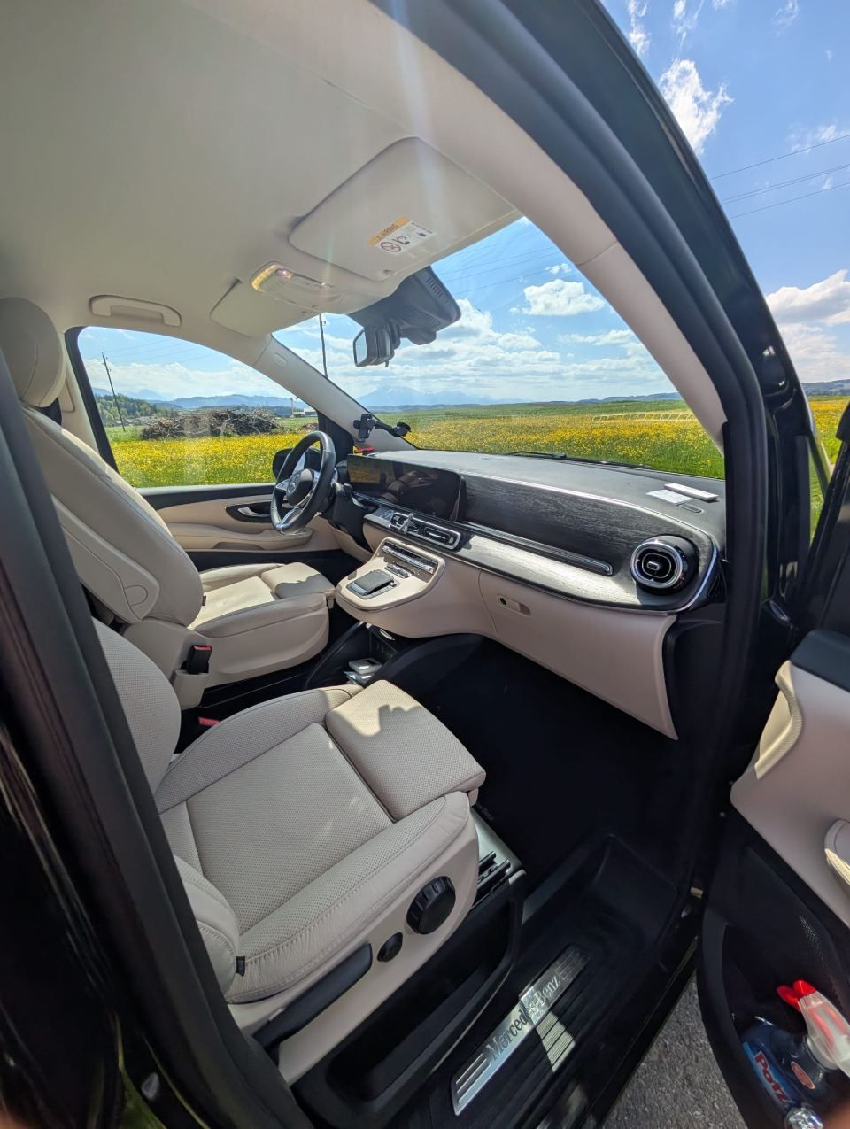 Der Innenraum des Wagens mit beigen Sitzen und Armaturenbrett, gesehen von der offenen Beifahrertür aus, bietet Blick auf ein sonniges Feld.
