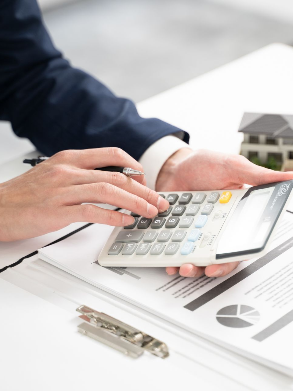 Une personne en costume utilise une calculatrice, des papiers et une maison miniature sur un bureau blanc.