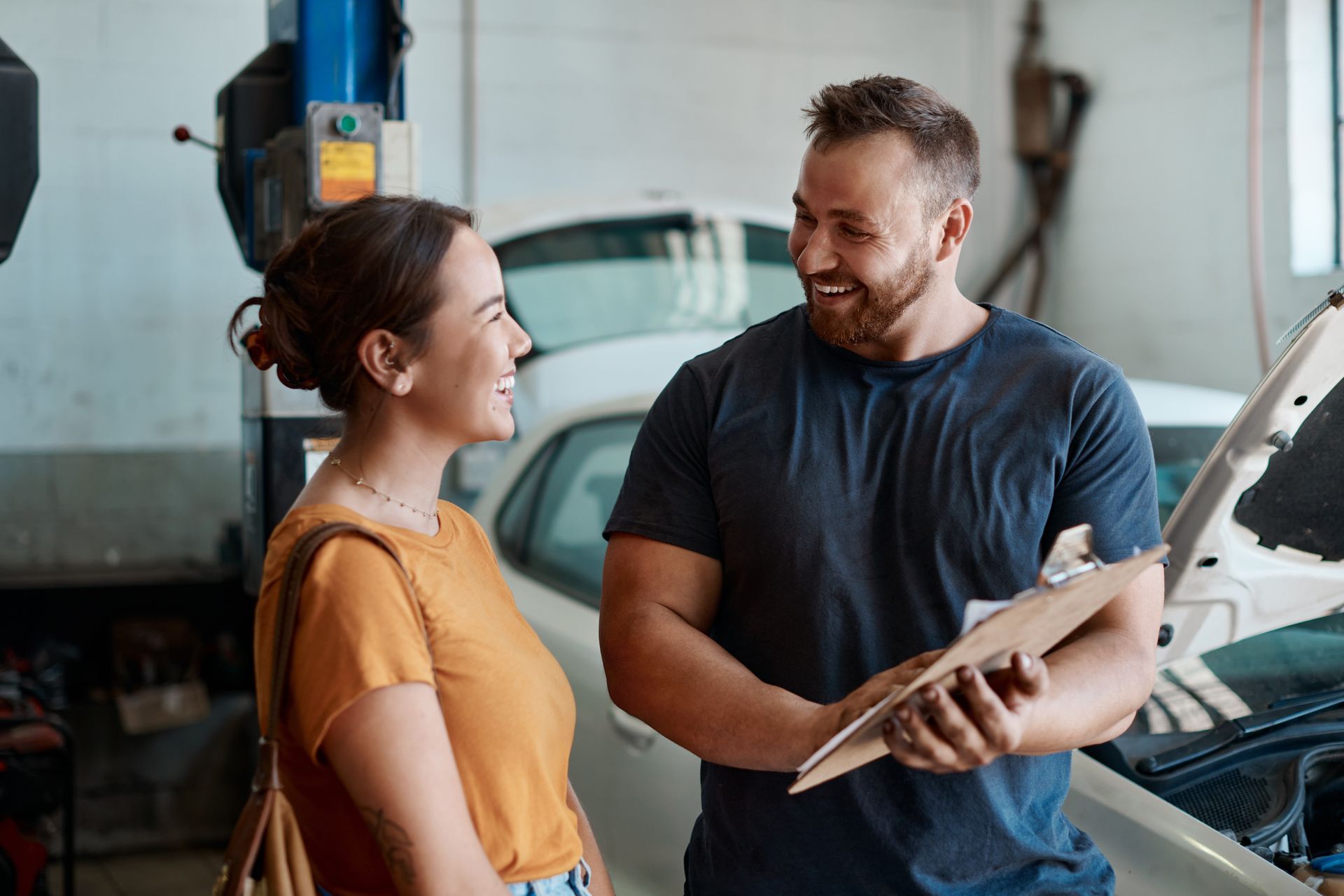 Une femme avec un haut orange discute avec un mécanicien dans un garage. Le mécanicien tient un bloc-notes et sourit. Une voiture est visible en arrière-plan.