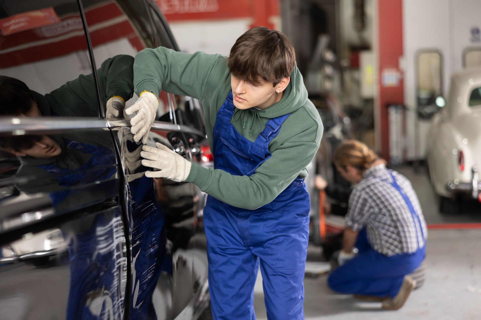 Un mécanicien en salopette et gants travaille sur une portière de voiture dans un garage.