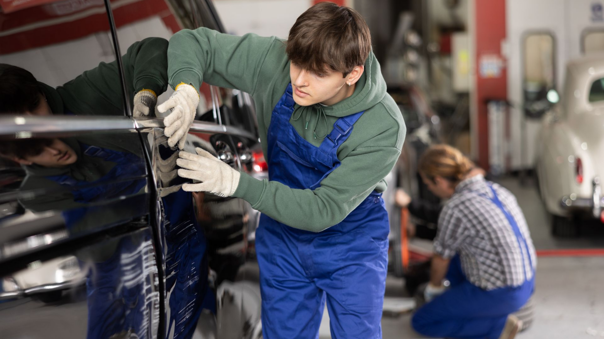 Un mécanicien en salopette bleue et sweat-shirt à capuche vert travaille sur une portière de voiture dans un garage.