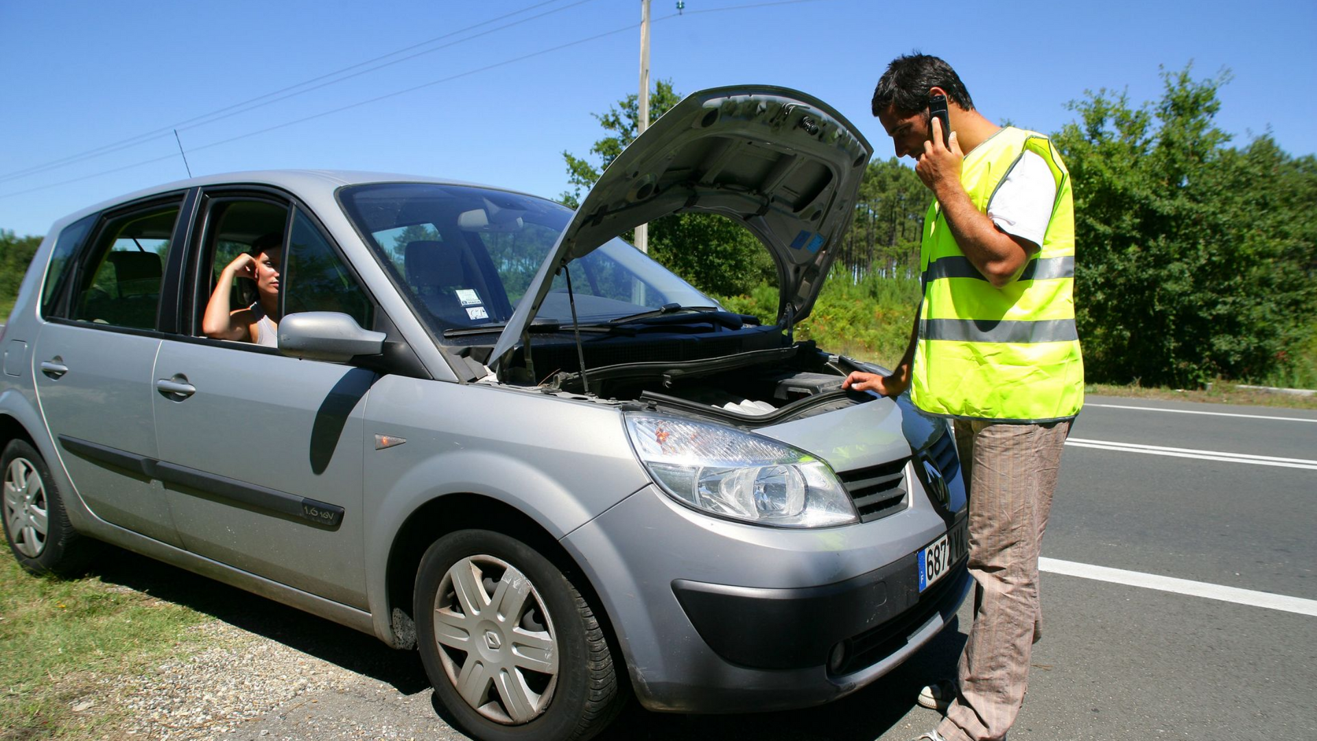Un homme en gilet jaune, au téléphone, se trouve près d'une voiture argentée en panne, capot ouvert, en bord de route.