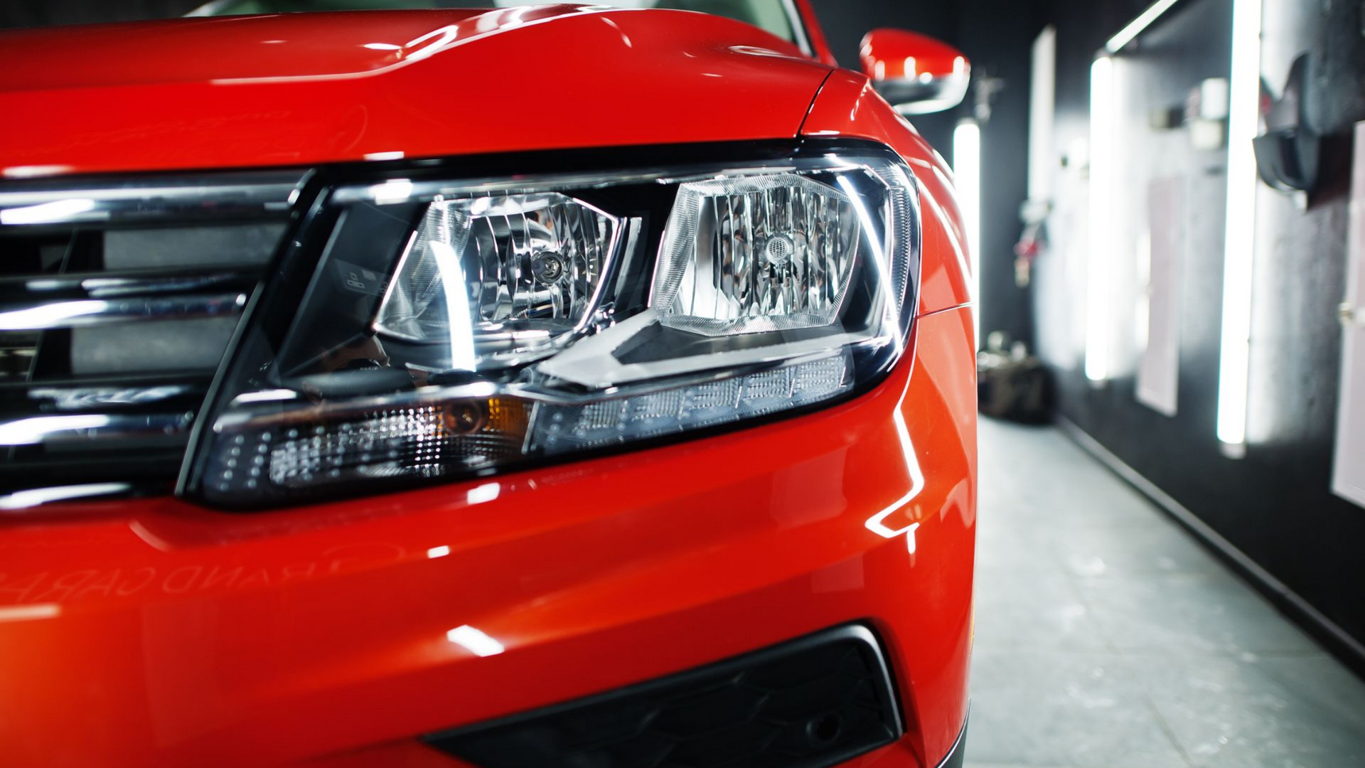 Gros plan sur le phare et le pare-chocs avant d'une voiture rouge vif, reflétant les lumières vives d'un garage.