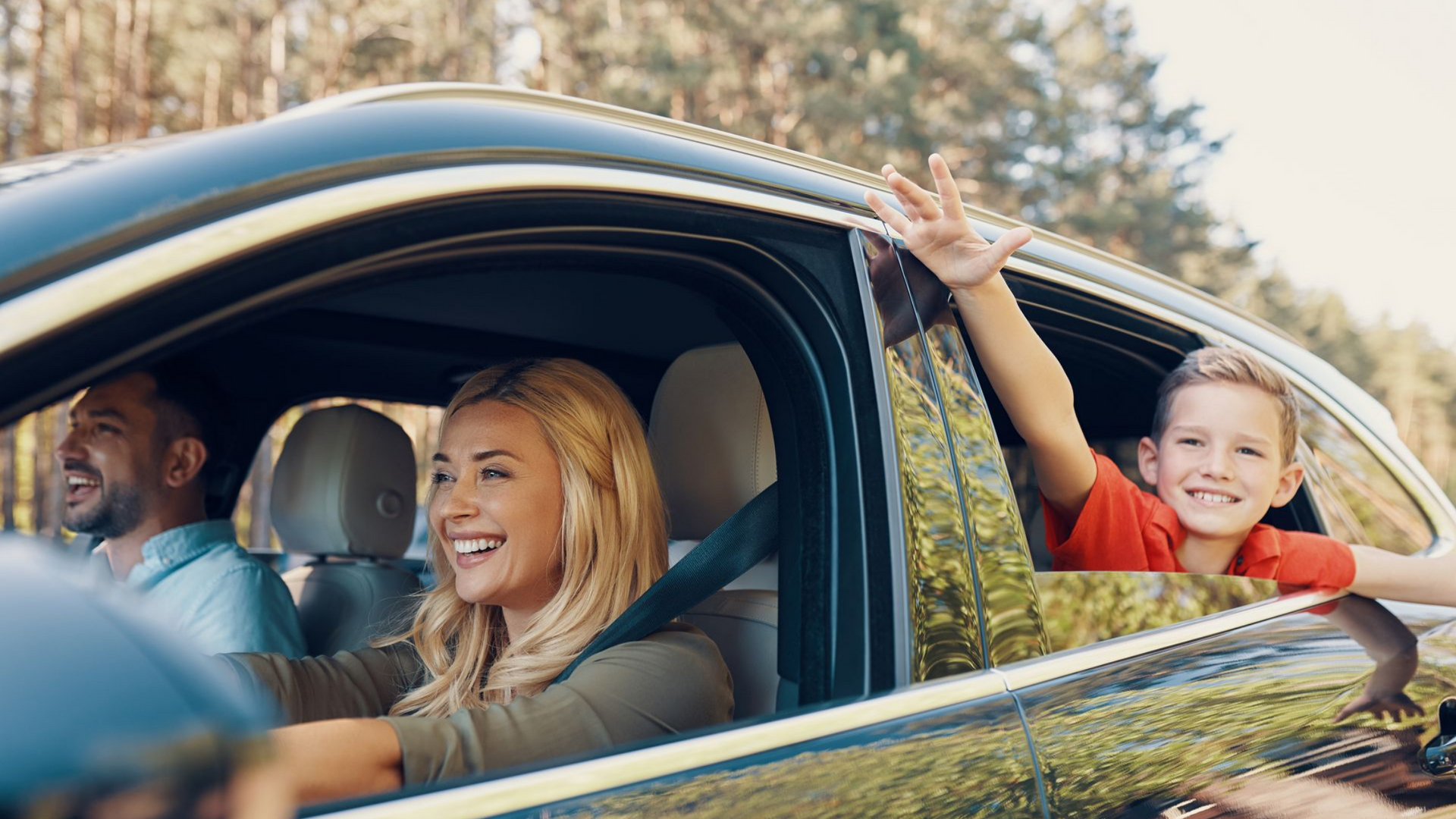 Famille en voiture : femme au volant, enfant faisant signe par la fenêtre, homme souriant. Forêt en arrière-plan.